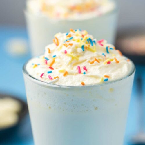 Tall glass of White Hot Chocolate with whipped cream and colorful sprinkles on top.