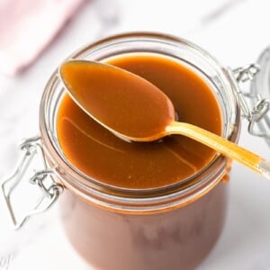 A spoon with caramel sauce on it sitting on the top of an open glass jar full of caramel sauce.