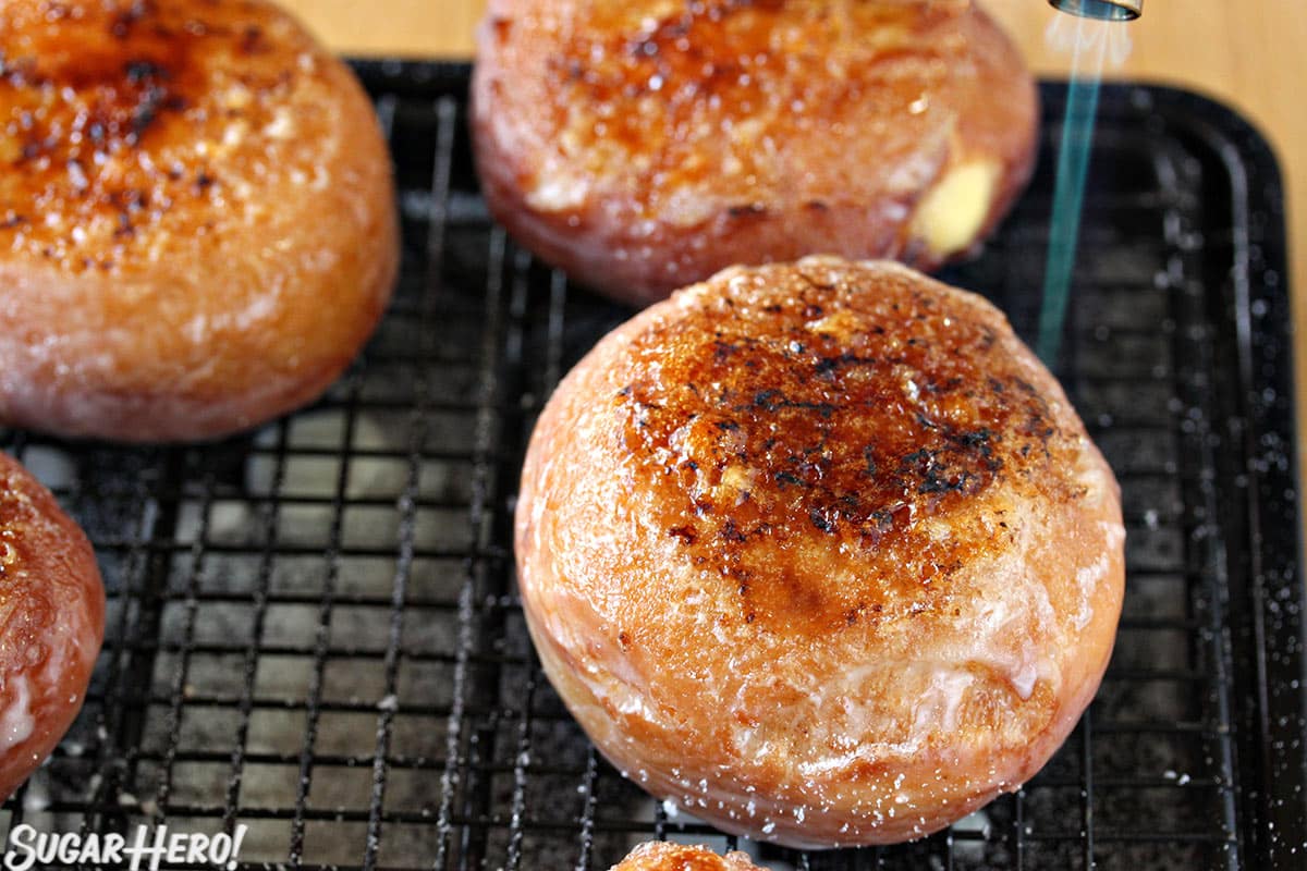 Donuts on a black wire rack, with a blowtorch caramelizing sugar on top of the doughnuts.