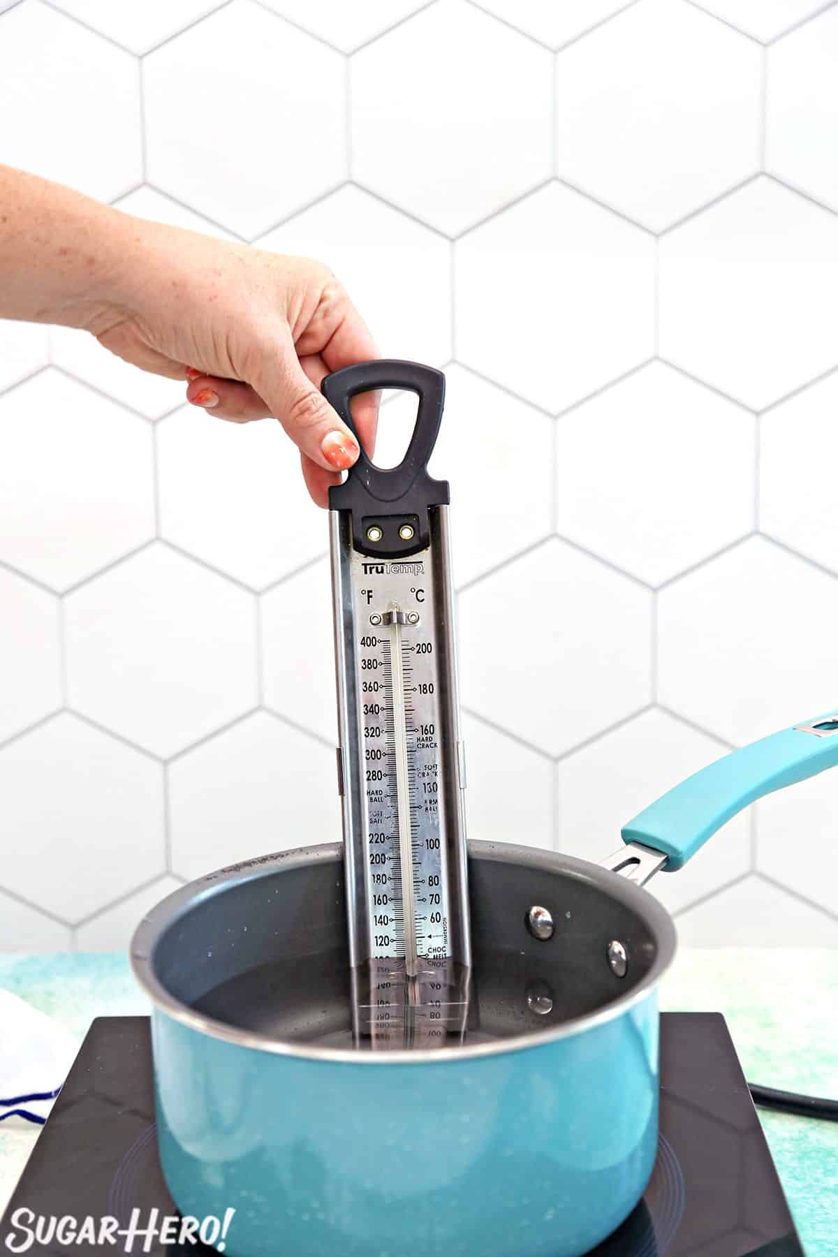 Hand inserting a thermometer in a a teal saucepan filled with water.