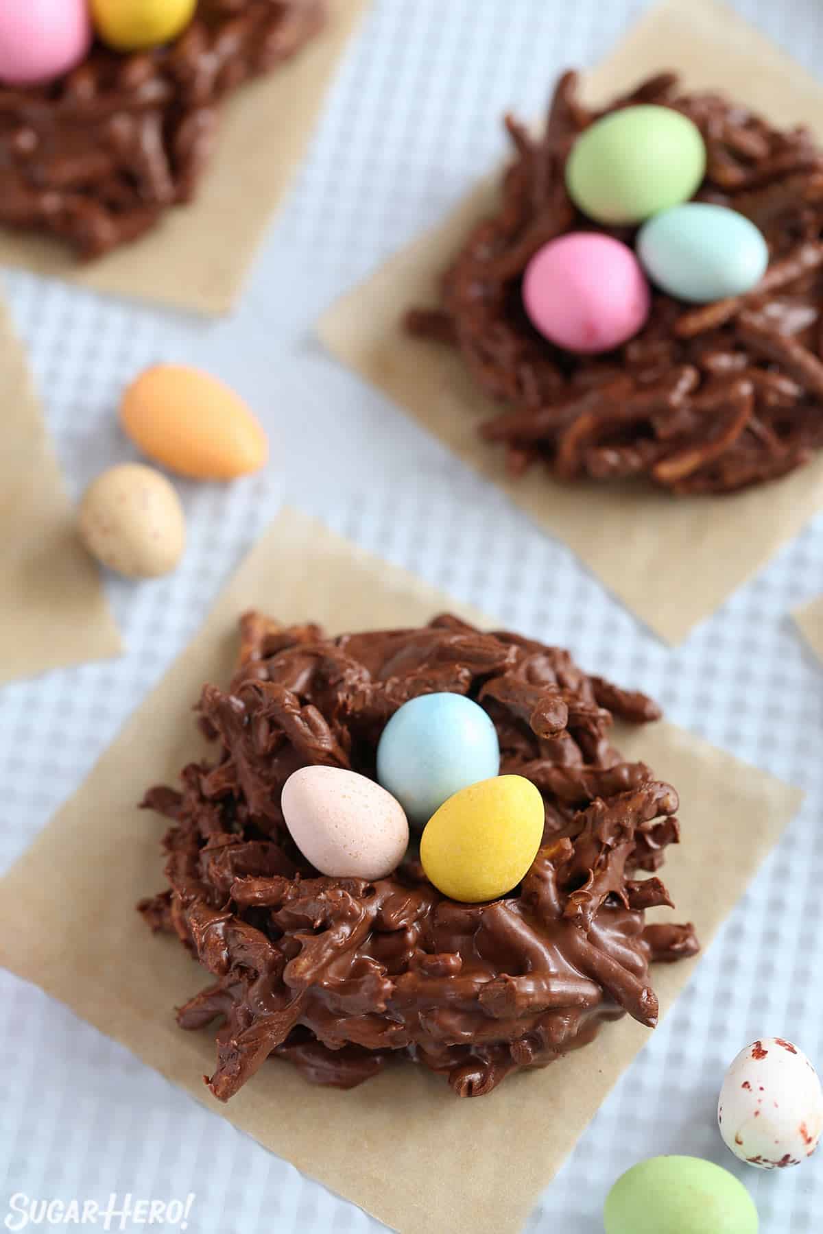 2 Chocolate Easter Nests on parchment squares with candy eggs scattered around.
