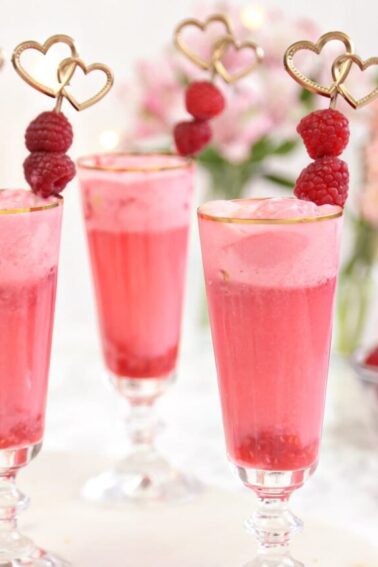 Four gold-rimmed glasses with pink raspberry punch and gold raspberry swizzle stick garnishes.