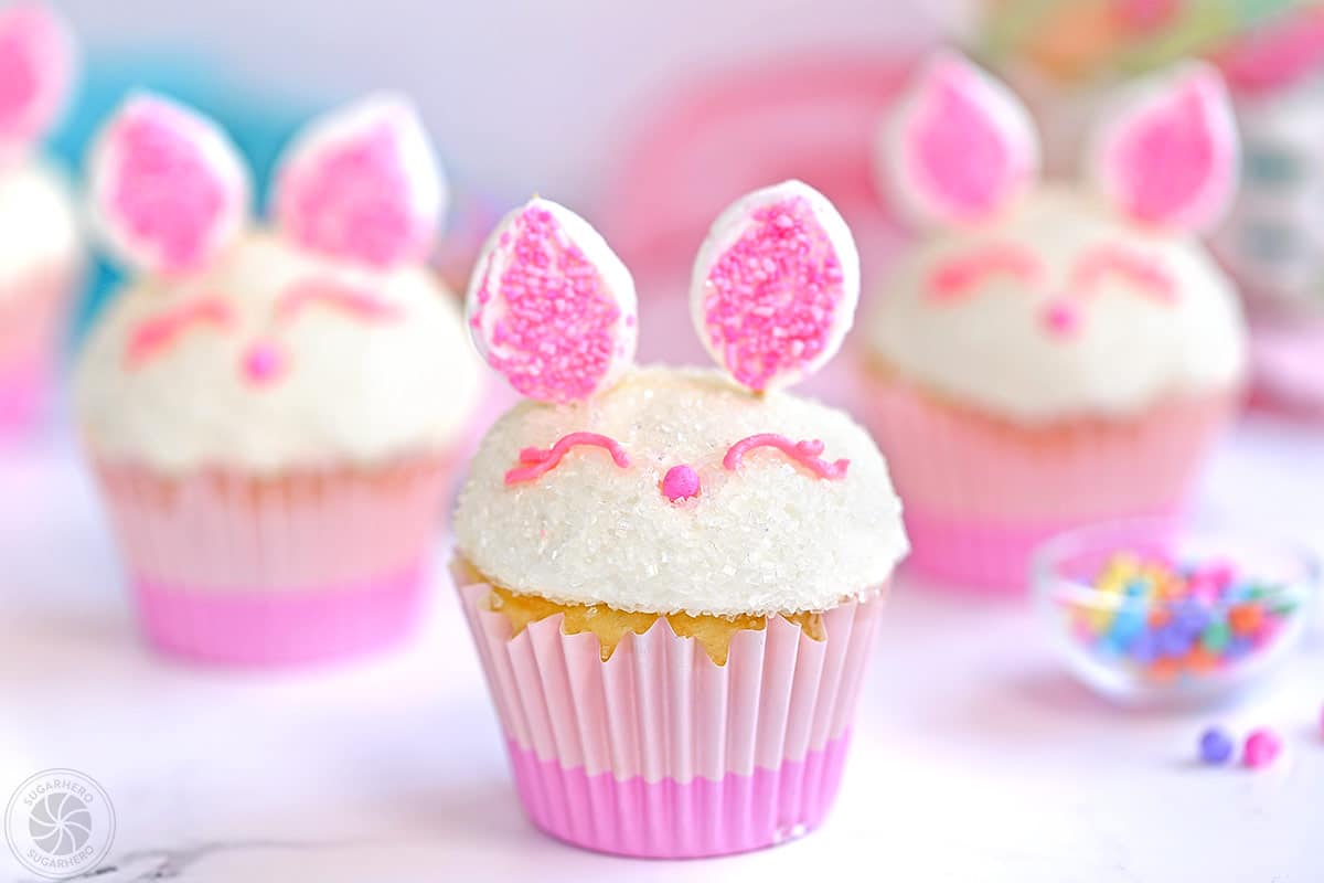 3 Easter Bunny Cupcakes on a white surface next to a bowl of sprinkles.