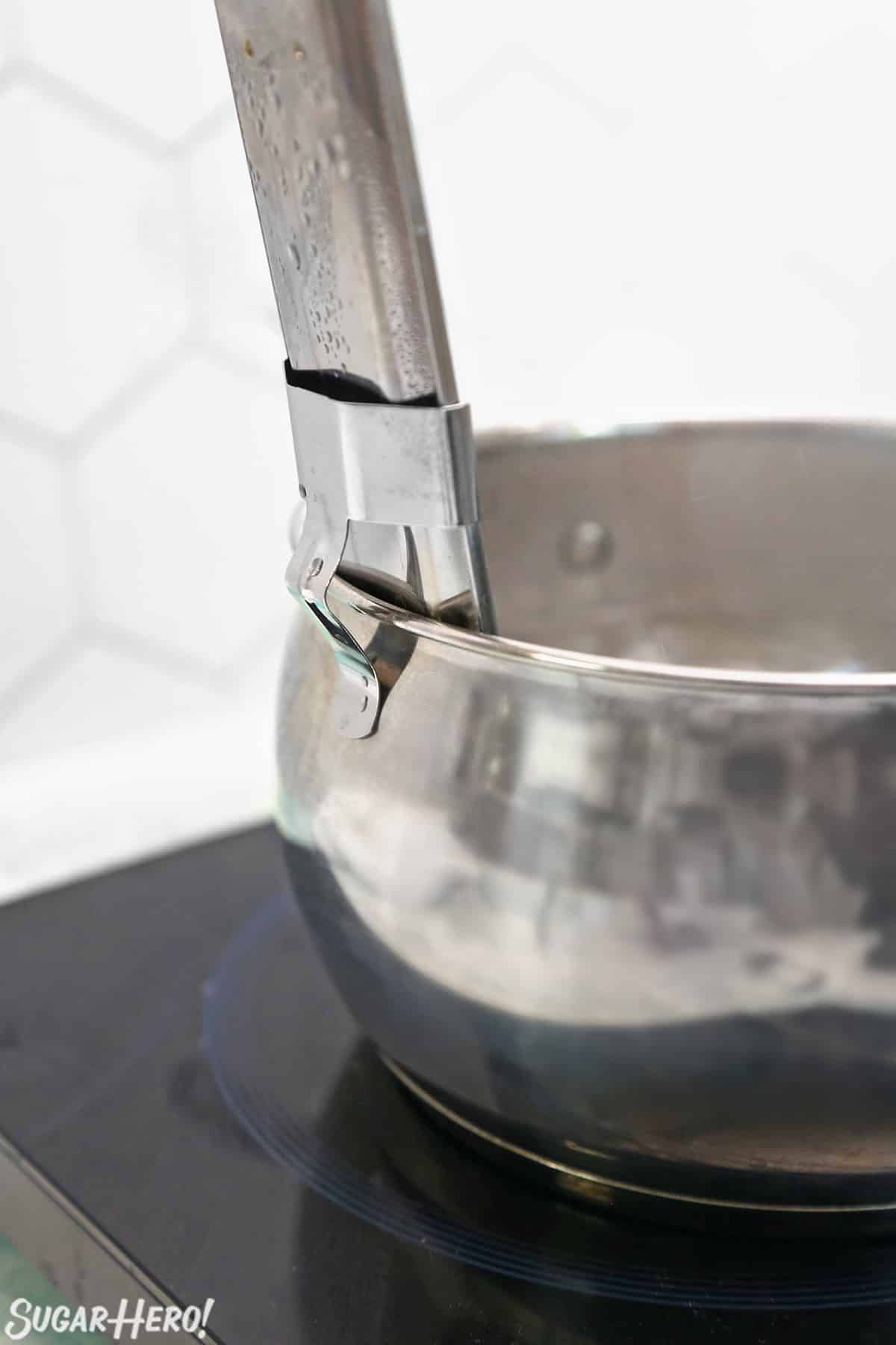 Side view of a metal candy thermometer clipped to the side of a metal pot.
