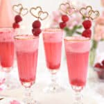 Four gold-rimmed glasses with pink raspberry punch and gold raspberry swizzle stick garnishes.