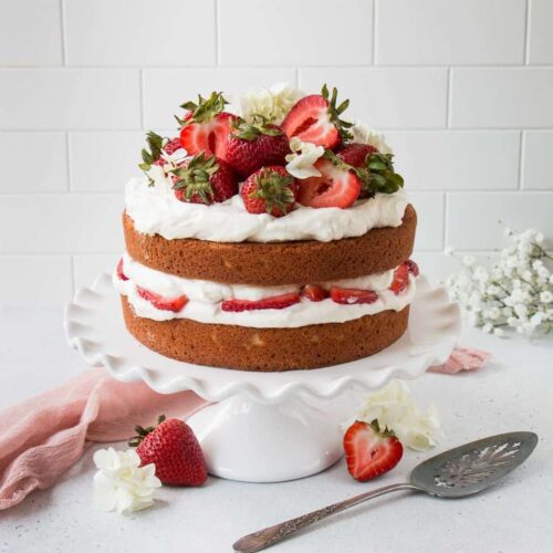 Front view of a layered Strawberry Shortcake Cake on a white cake platter surrounded by strawberries and white flowers.