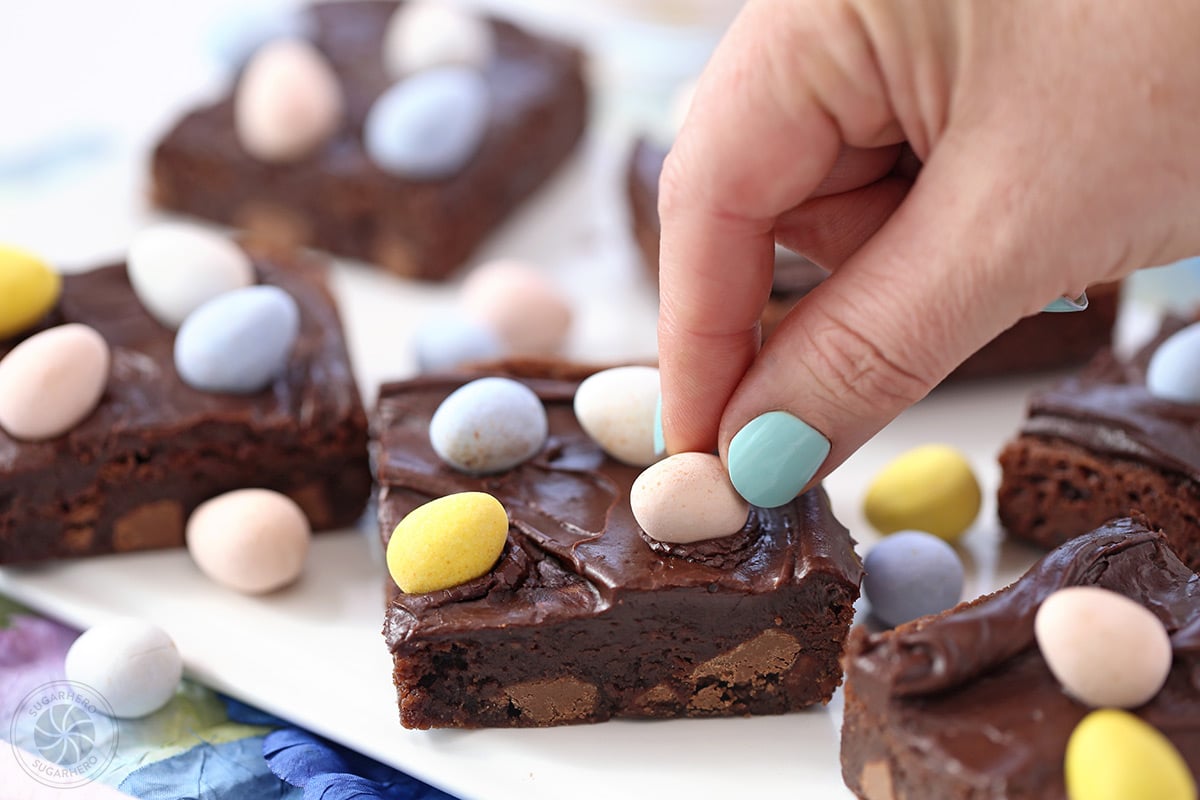 Hand with blue fingernails pressing a chocolate egg into the frosting on top of a brownie.