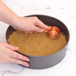 Using a copper measuring cup to press graham cracker crust into the base of a springform pan.