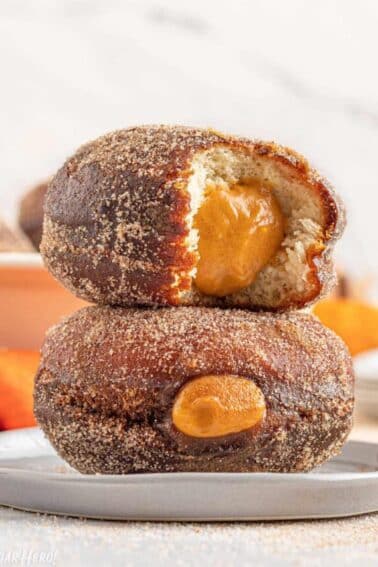 2 Pumpkin Cream Filled Donuts on a small gray plate stacked one on top of the other and a bite removed from one to show the pumpkin filling.