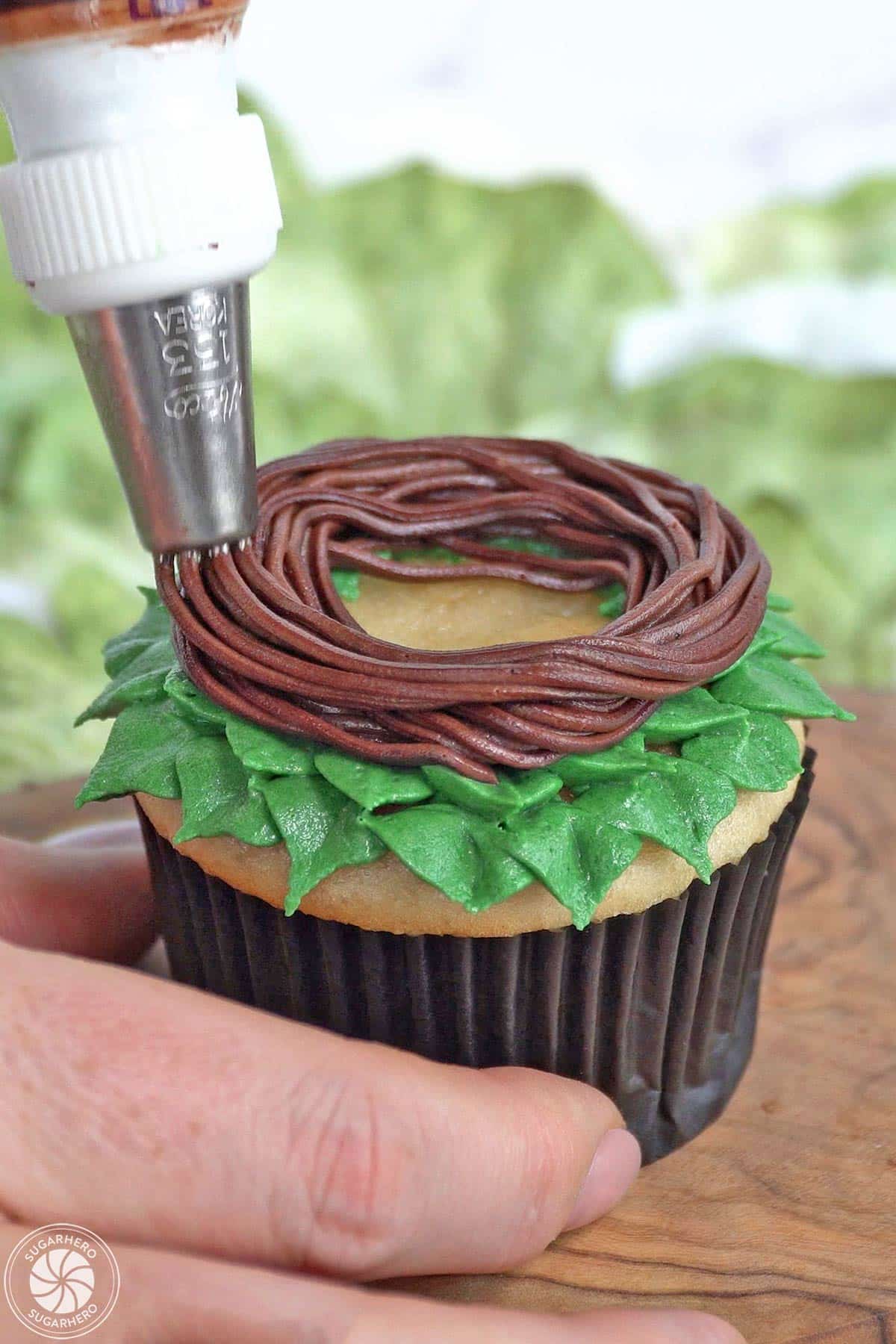 Piping a chocolate buttercream nest around the edge of a cupcake.