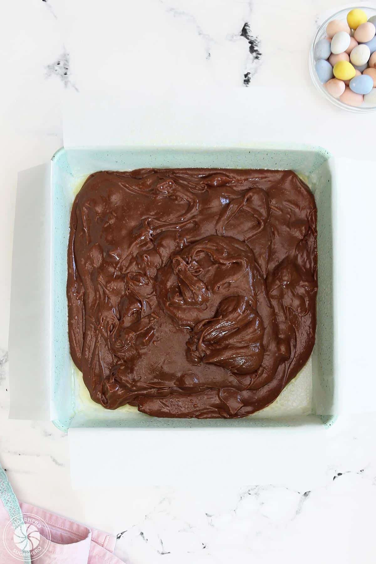 Overhead shot of brownie batter in a green square pan, on a white countertop.