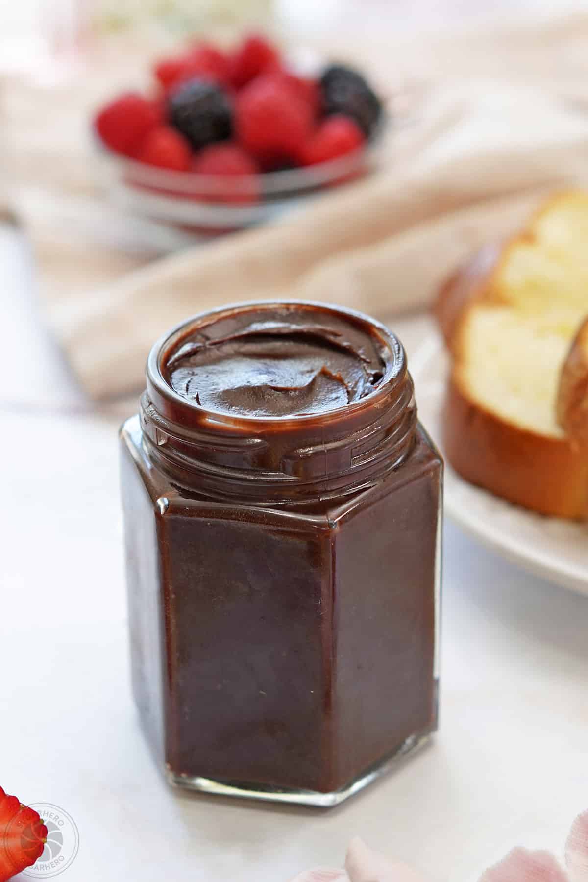 A glass jar of Chocolate Spread next to fruit and bread.