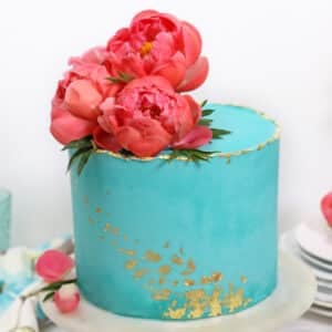 Teal cake with edible gold leaf accents and pink peonies on top.