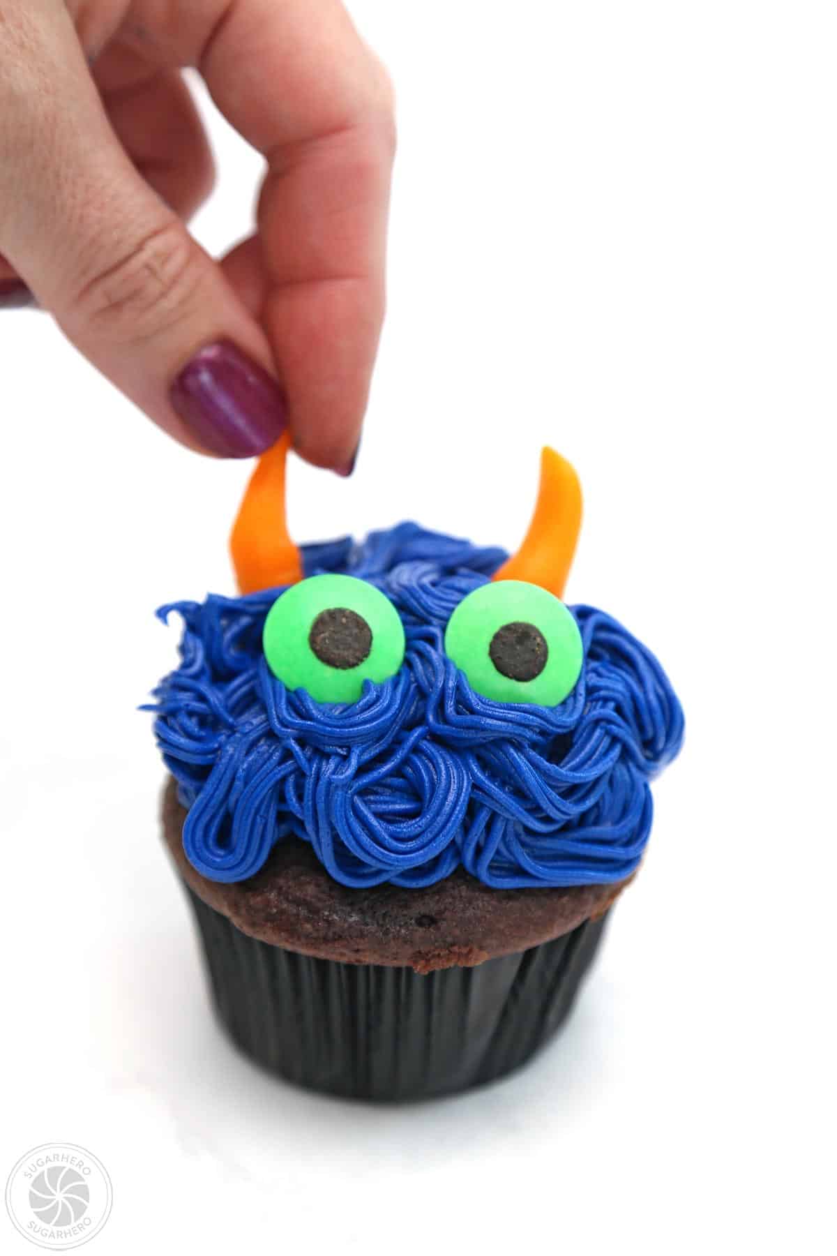 Hand placing horns on a blue monster cupcake.