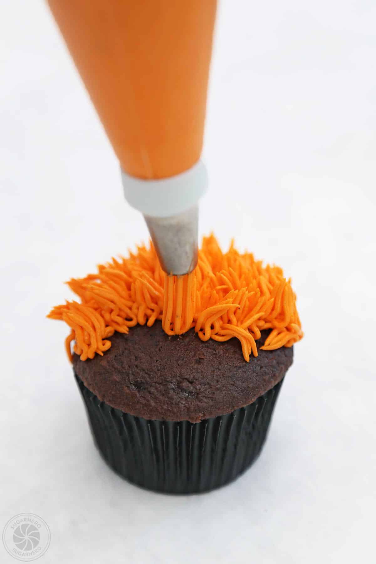 Using a grass tip to pipe orange frosting hair on a monster cupcake.