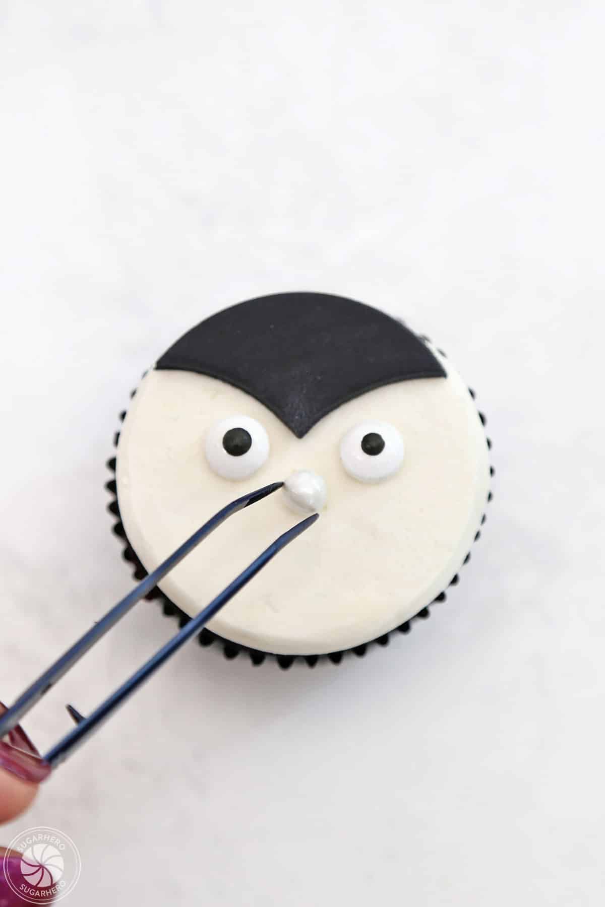 Hand using tweezers to place a candy nose on a vampire cupcake.