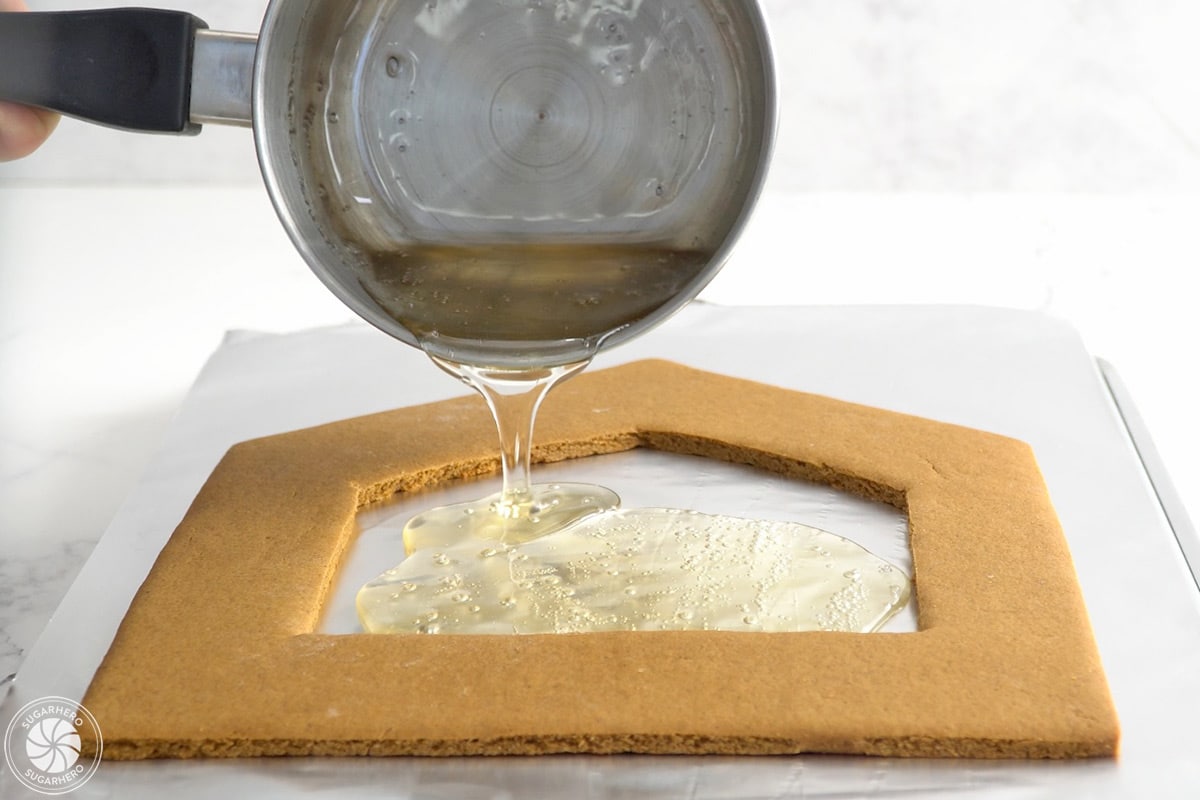 Hand pouring sugar syrup into a gingerbread house panel for the window.