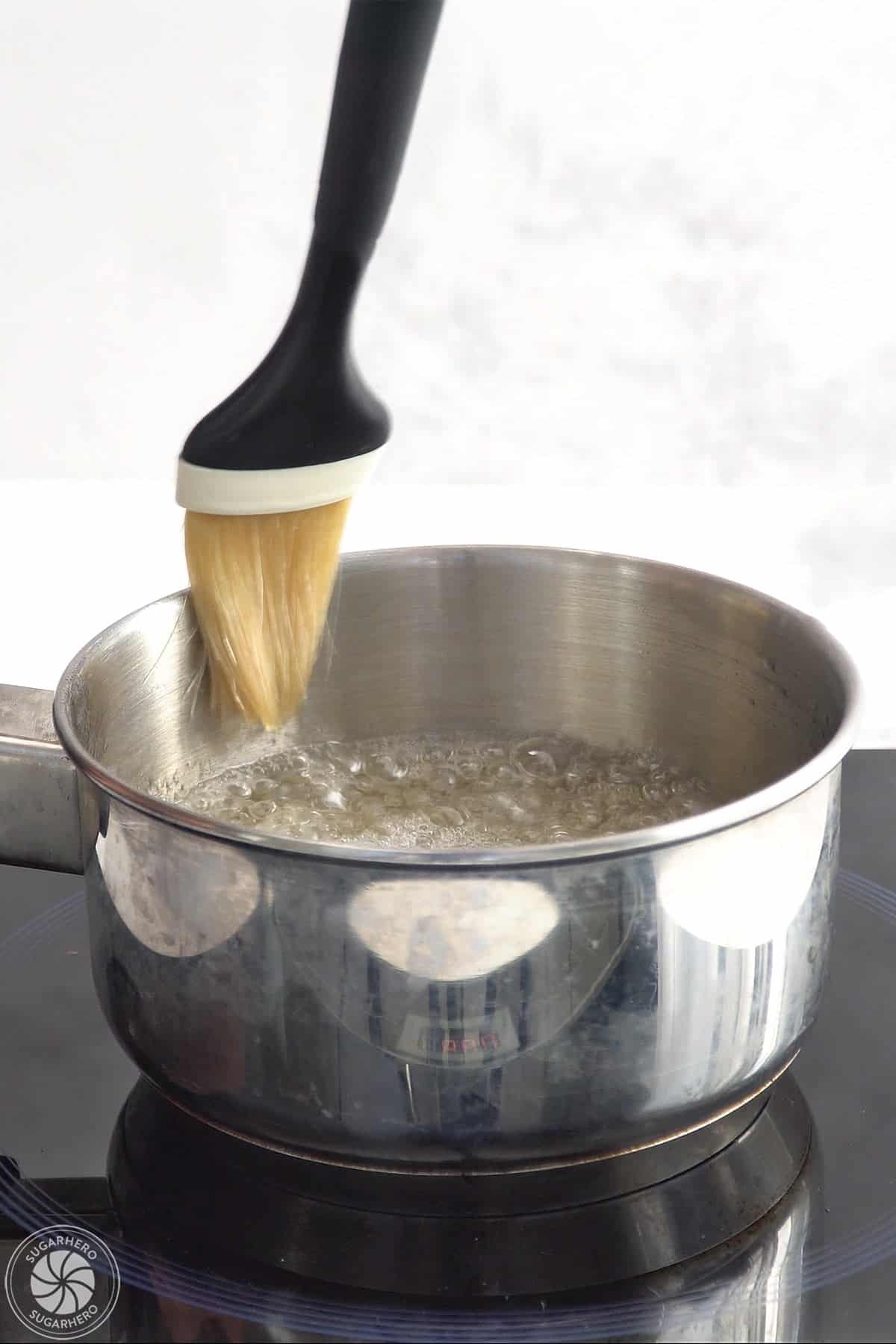 Using a pastry brush to wipe away sugar crystals around the rim of a pot of boiling sugar syrup.