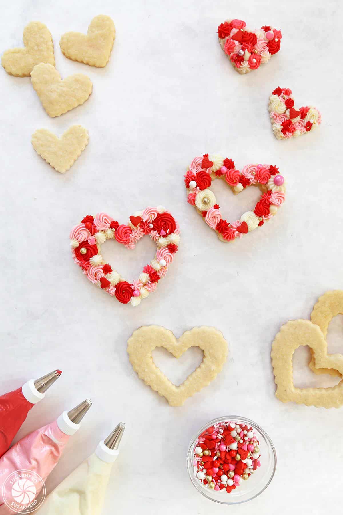 Unfrosted sugar cookies next to decorated cookies, a bowl of sprinkles, and 3 piping bags filled with red, pink and white frosting.
