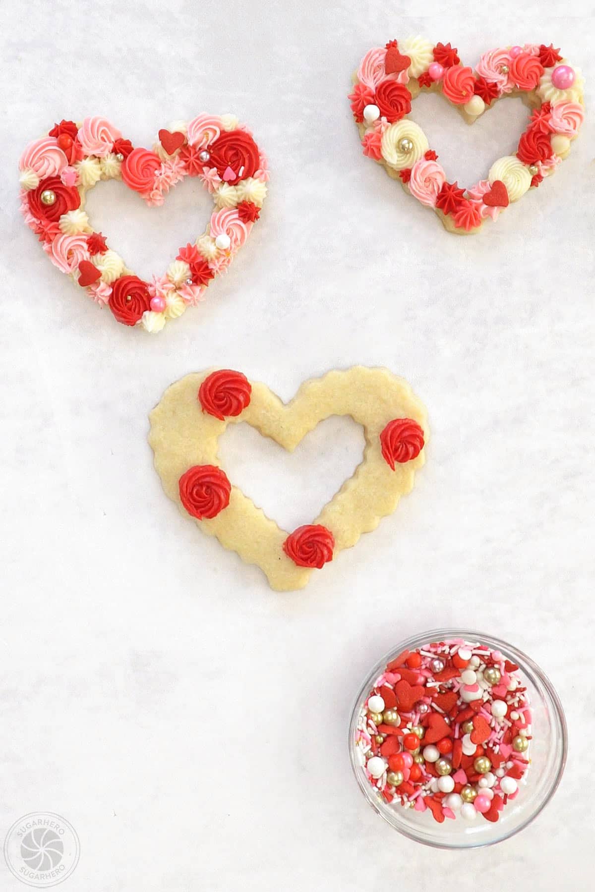 Red frosting swirled into tiny rosettes on a sugar cookie next to two decorated sugar cookies and a bowl of sprinkles.