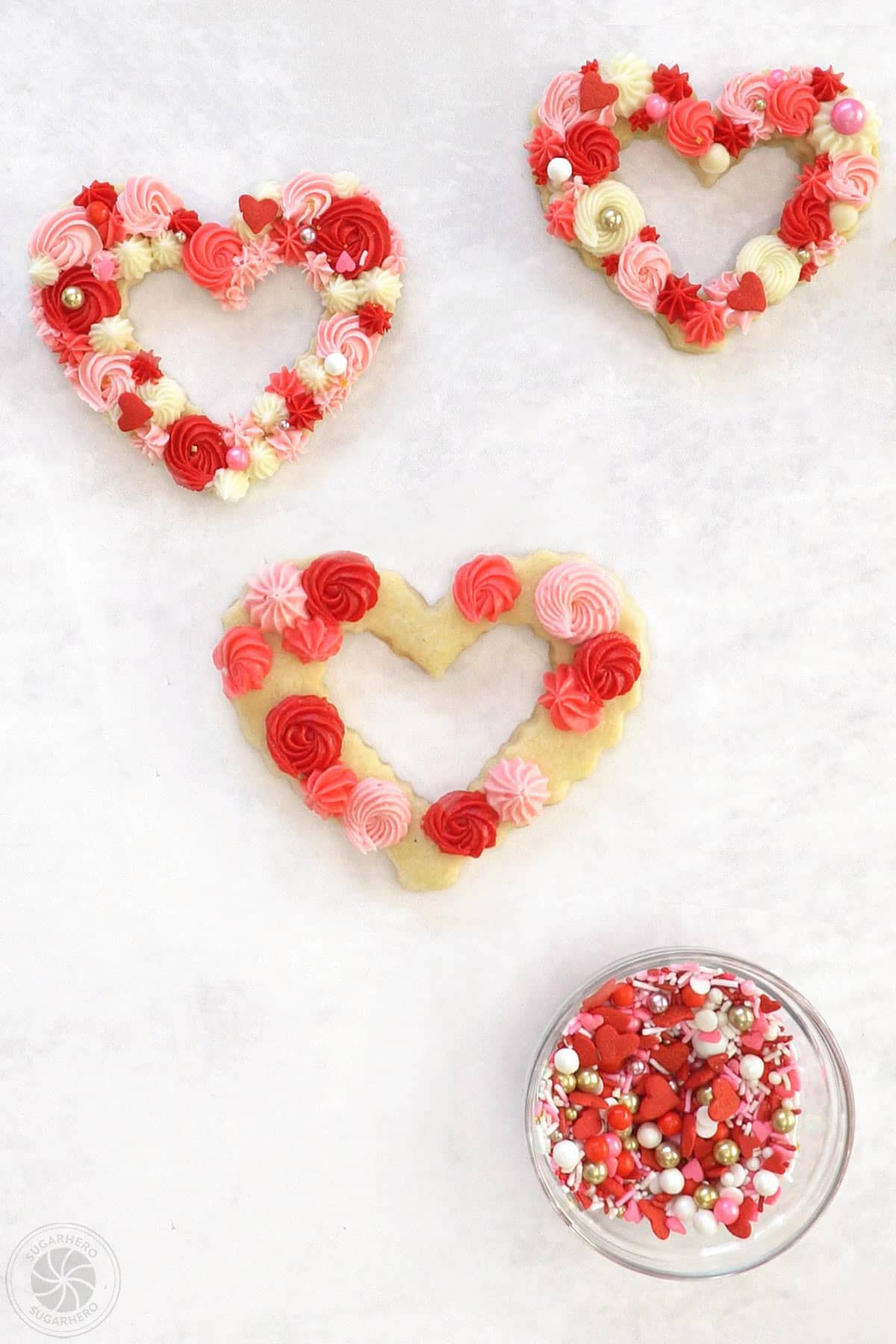 Two decorated sugar cookies next to a cookie that is being decorated with red and pink rosettes and swirls next to a bowl of sprinkles.