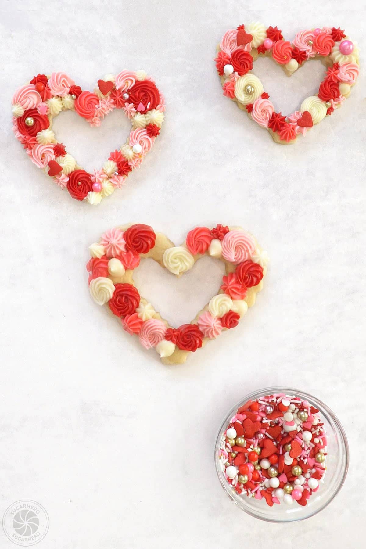 Two decorated sugar cookies next to a cookie that is being decorated with red, pink and white rosettes and swirls next to a bowl of sprinkles.