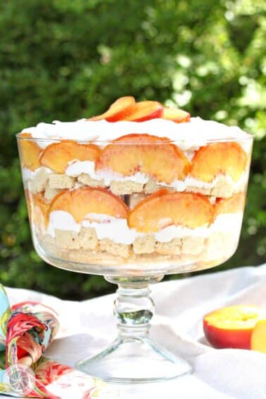 A peaches and cream trifle on a white table cloth with greenery in the background.
